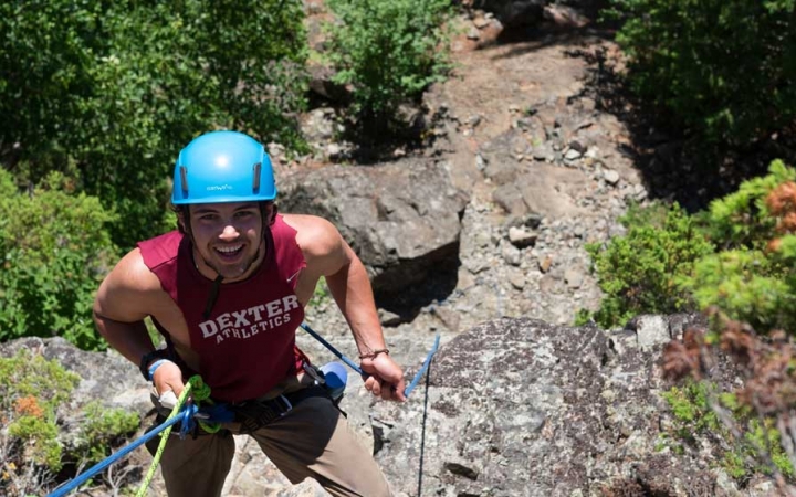 A person wearing safety gear is secured by ropes as they look up at the camera and smile while rock climbing.